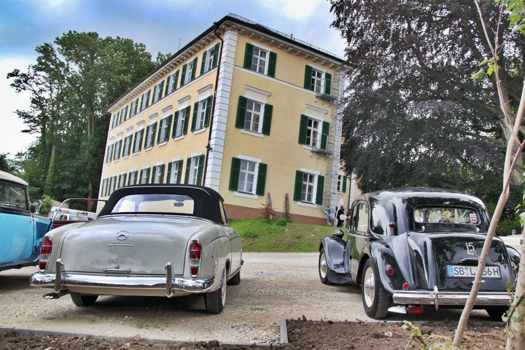 Eine Oldtimerausstellung vor einem großen, gelben, dreistöckigen Gebäude mit grünen Fensterläden. Die Szene umfasst klassische Autos, darunter ein silbernes Mercedes-Cabrio und eine schwarze Limousine, die auf einem von Bäumen gesäumten Kiesweg geparkt sind. Hotel Schloss Burgellern