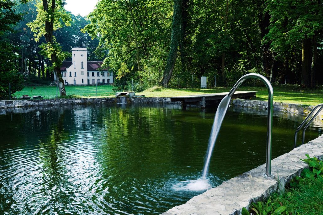 Ein ruhiger Teich mit Steinrand und Metallhandlauf verfügt über einen kaskadenförmigen Wasserspeier. Er liegt inmitten eines üppigen grünen Parks und im Hintergrund ist ein weißes Gebäude mit rotem Dach zu sehen, umgeben von hohen Bäumen. Hotel Schloss Burgellern