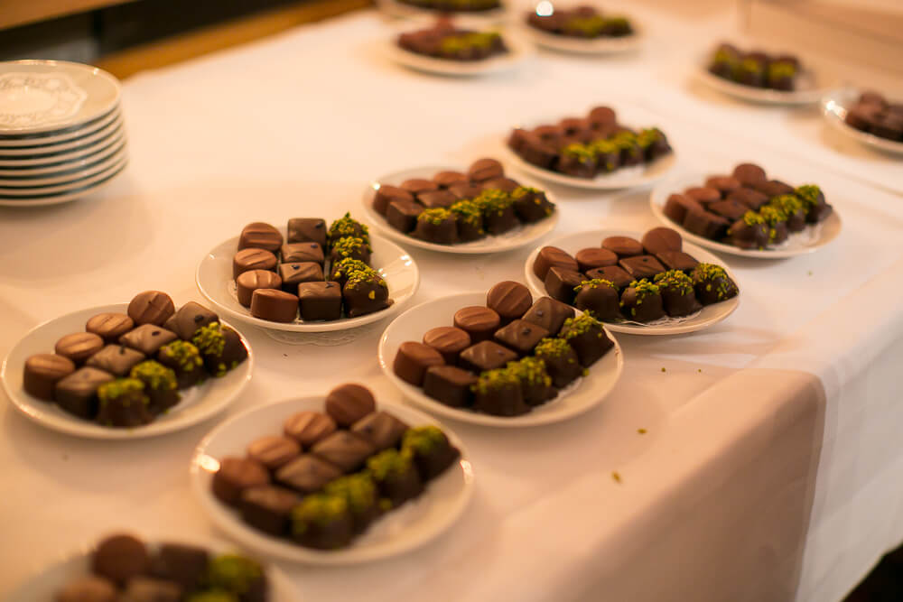 Auf einer weißen Tischdecke angeordnete Teller, jeder mit verschiedenen Pralinen gefüllt und mit gehackten grünen Pistazien garniert. Im Hintergrund sind gestapelte weiße Teller zu sehen. Hotel Schloss Burgellern