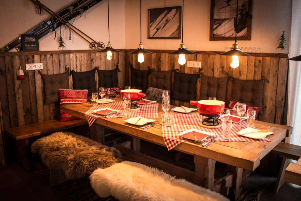 Ein gemütlicher Essbereich mit einem für eine Mahlzeit gedeckten Holztisch mit rotkarierten Tischdecken, Fonduetöpfen und Weingläsern. Der Raum ist mit rustikalen Holzvertäfelungen, Vintage-Skiern an der Wand und warmen Hängelampen ausgestattet. Hotel Schloss Burgellern