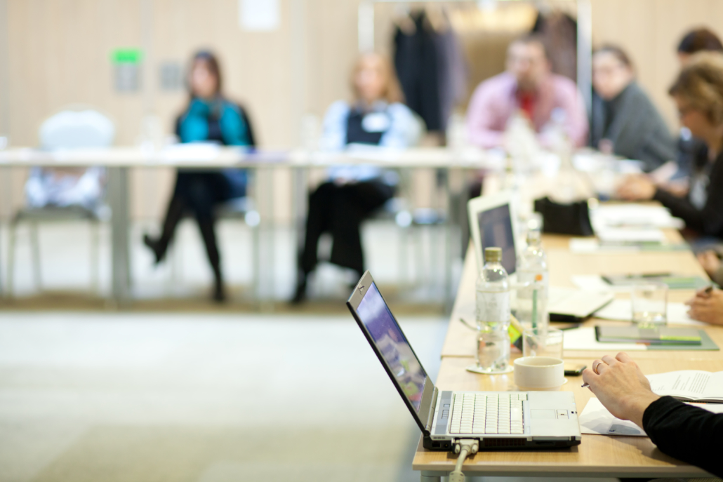 Ein Besprechungsraum mit Menschen, die um einen langen Konferenztisch sitzen. Im Vordergrund sind ein Laptop und ein Notizblock auf dem Tisch zu sehen. Im Hintergrund sind verschwommen Teilnehmer zu sehen, die aufmerksam zuhören. Hotel Schloss Burgellern