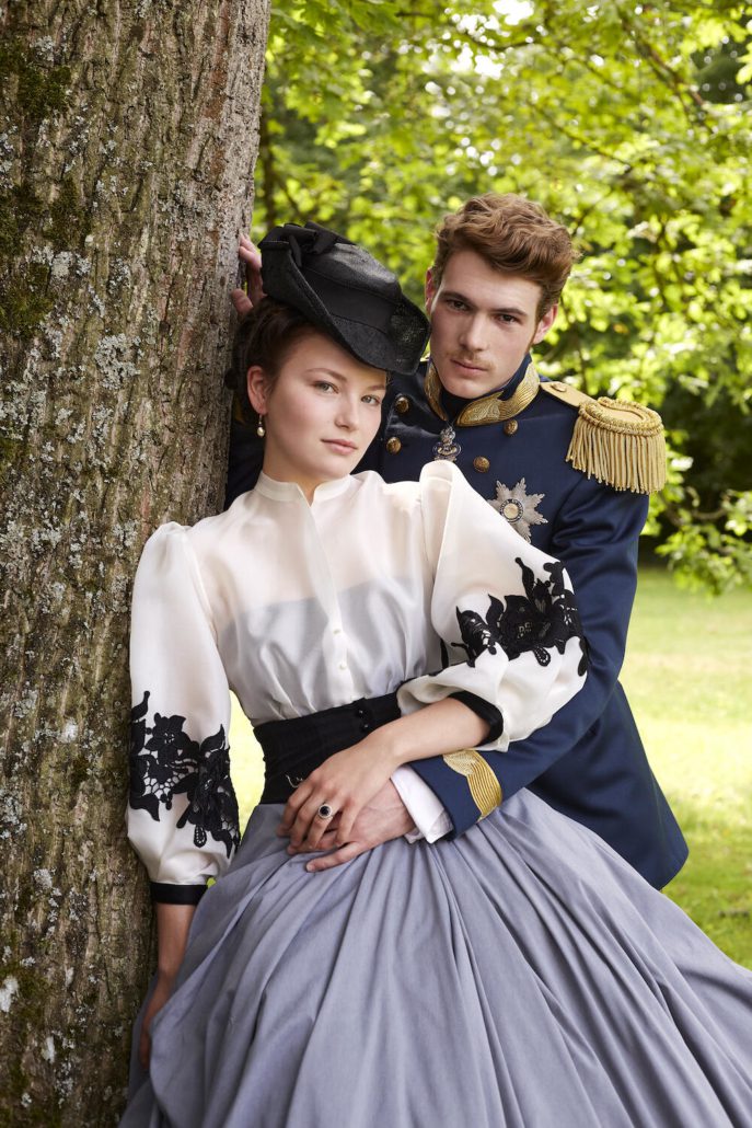 Ein Paar in viktorianischer Kleidung posiert im Freien inmitten der üppigen grünen Vegetation Bambergs. Die Frau in einer weißen Bluse mit schwarzer Spitze und einem grauen Rock lehnt an einem Baum. Der Mann in einer blauen Militäruniform mit goldenen Akzenten steht hinter ihr und fängt den Charme der historischen Stadt ein. Hotel Schloss Burgellern