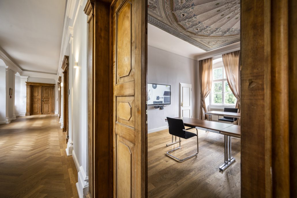 Ein geräumiger Raum mit Holzwänden und -boden, der sich perfekt für jeden Tagungsplaner eignet, verfügt über einen großen Tisch und Stühle. Durch ein Fenster mit Vorhängen fällt sanftes Licht in den Raum. Die aufwendig gestaltete Decke ergänzt den an einer Wand angebrachten Flachbildfernseher. Hotel Schloss Burgellern